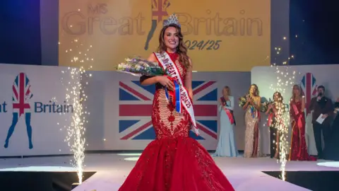 The Perfect Image Emma on stage is a red ballroom dress, with a MS GB 2025 sash in white and a diamond tiara. she's holding a bouquet of flowers. There are fireworks going off and you can see other contestants in their gowns in the background 