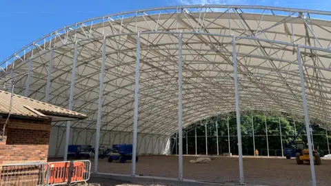 A construction site with a curved, metal framed roof over some flat ground with construction vehicles inside