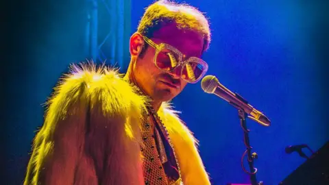 Richard Reeves Richard is dressed as Elton in a fur coat standing by a microphone on stage.