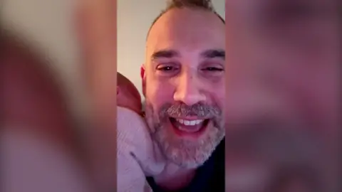 A man smiling to camera, with his face visible and a newborn baby on his shoulder.