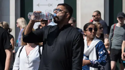 A man in a London street on a sunny day takes a drink of water from a plastic bottle. 