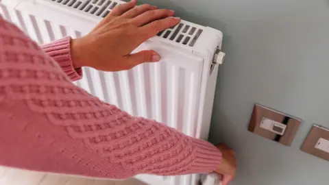 A woman wearing a pink woolly jumper is touching a radiator with her left hand while adjusting the knob on the radiator with her right hand, 