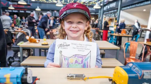 Paul Campbell Rosie is wearing a claret train driver's cap. She is holding up her winning drawing. She is at a table with a Thomas the Tank Engine loco and a yellow and blue diesel locomotive. There are other tables, and people, behind her in a food hall of the Victorian Market.