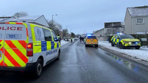 Police vehicles, including a van and a SUV parked on the street in Inverness. The road is wet while the pavements are covered in a layer of snow. There are rows of houses along the road.