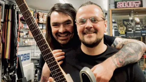 Sam Orr is smiling as he stands behind Mark Wood in Sam's guitar shop. Mark Wood is also smiling as he holds his guitar, with the neck of the guitar and the frets visible.