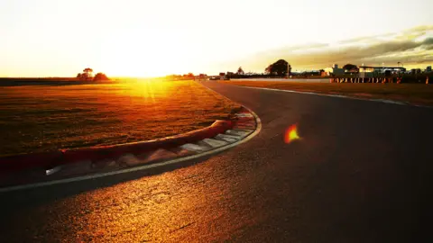 Motorsport UK Snetterton racetrack, with the sun in the distance, you can see a track, grass area and a number of buildings in the distance, to the right. 