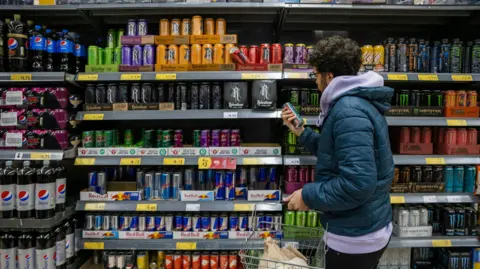 Getty Images A young man, dressed in a puffer coat, shops in a supermarket, picking up a can of energy drink and reading the nutritional information printed on the back