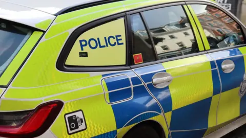 A police car. The livery is fluorescent yellow and blue checks and there is a sticker of a speed camera near the petrol cap and the word POLICE printed on the back window. There are reflections of buildings in the windows.  