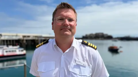 BBC James Way is wearing his uniform, complete with epaulets, and is standing in front of the harbour in the sunshine. He has short dark blonde hair and is wearing apair of glasses.