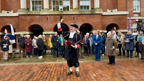 Chichester Women's History Group A person dressed in historic clothing holds up a handbell in front of a crowd outside a red‑brick building with arched entrances.