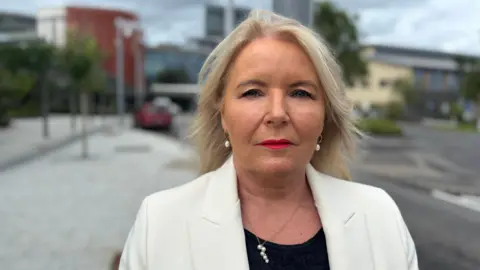 Pat Cullen looking at the camera. She is standing outside wearing a white blazer, black top, a pearl necklace and earrings and red lipstick. She has long blonde hair.