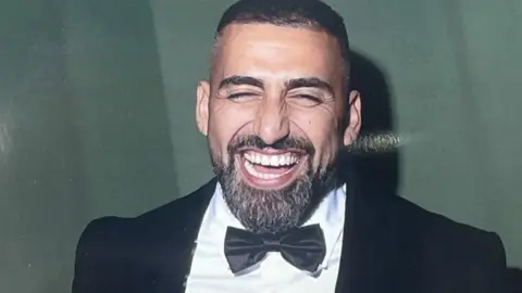Sina Ghami in a black tuxedo, white shirt and black bowtie. He has short dark hair, a dark beard with a few grey hairs. He is laughing and his eyes are almost full closed.