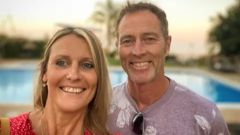 PA Media Lindsay, left, who has blonde mid length hair and is wearing a red dress, next to her husband Craig, who is wearing a mauve t-shirt with a white print. Both are smiling in the sun in front of a blue pool. 