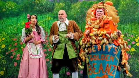 BBC Belle, her father and panto dame Betty Bon Bon on stage at the Lawrence Batley Theatre with a castle meadow backdrop in the background. The panto dame is wearing a brightly coloured costume which looks like a tin of baked beans has been poured over them.