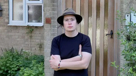Josiah English is standing in front of a guesthouse and is looking directly at the camera with his arms crossed. He is wearing a black T-shirt and a hat.