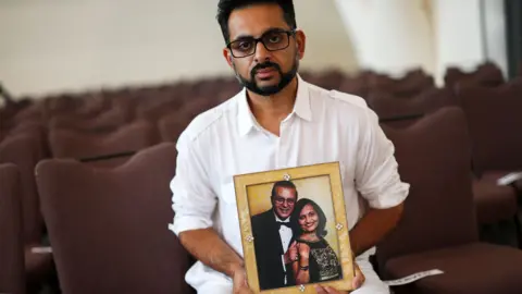 A photo of an Indian man wearing a white shirt holding a framed photograph of his mother