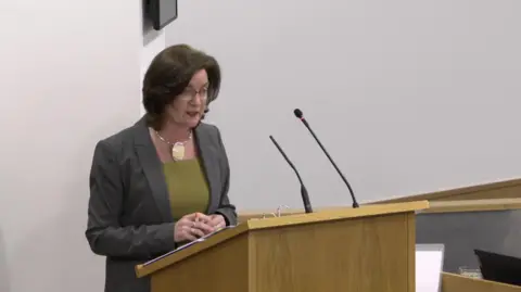 Senedd Cymru Eluned Morgan standing behind a light coloured wooden lectern in the temporary Senedd chamber in Cardiff Bay.