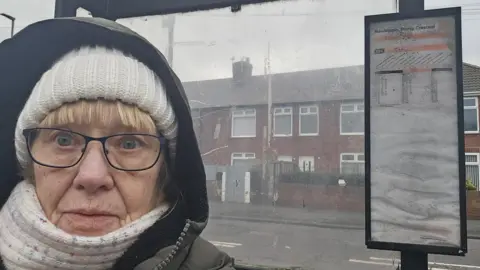 Liz Hobson is standing in a bus stop with a thick white hat on and white scarf and a thick coat on with the hood pulled up. She has a blonde fringe and wears black-rimmed spectacles. There is a bus timetable over her left shoulder. A row of two-stoery terraced houses are on the other side of the road, behind the clear Perspex shelter windows. 