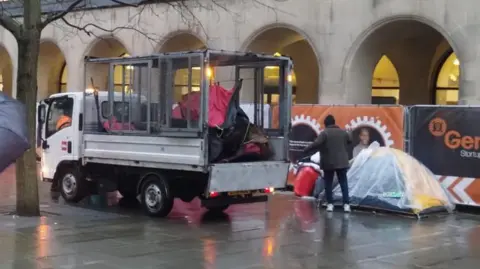 A small van parked in front of the arches at St Peter's Square as council workers throw red tents into a trailer on the back. A man in an umbrella is stood watching the scene. 