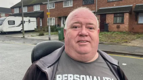 A white haired man sits on a mobility scooter in a t-shirt and hooded jumper, in front of a row of houses, one of which has a white caravan parked out front.