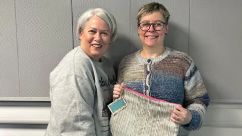 BBC A woman with short grey hair and wearing a grey jumper, and a woman with short blonde hair, glasses and a multicoloured stripy cardigan. The woman wearing glasses is holding up a beige bag with a pink zip and a ribbon logo on the front. They are both smiling at the camera.