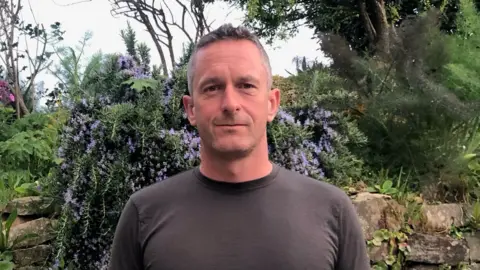 Tom Whitmore is standing outside. He has short grey-brown hair and looks straight at the camera. Behind him is a green plant with purple flowers on it, a stone wall and a few trees in the background. He is wearing a grey t-shirt.