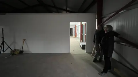ELLEN KNIGHT/BBC Peter and Alison inside the shell of the building, looking up at the ceiling. It's fairly dark inside, but a room beyond is lit up using floodlights. The walls are white corrugated metal with dark brown beams. The floor is made of concrete.