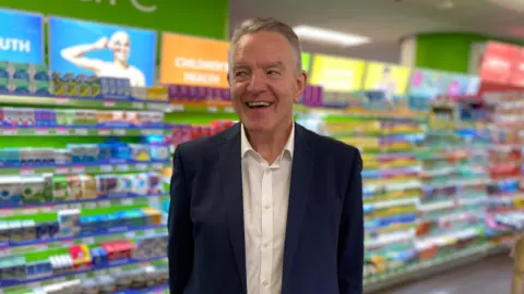Peter Macnab, Superdrug's CEO is standing in the Guernsey store wearing a white shirt and a navy blue suit jacket. He has short, well kept very light brown hair.