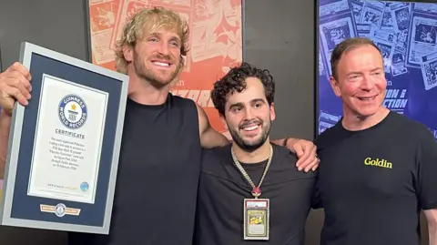 Logan Paul holding up a framed Guinness World Records certificate, with his arm around AJ Scaramucci, who is wearing the card around his neck. Ken Goldin is next to them, and they're all smiling for the cameras