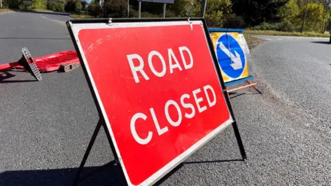 Red road closed sign in front of red plastic barriers which have fallen over onto the road