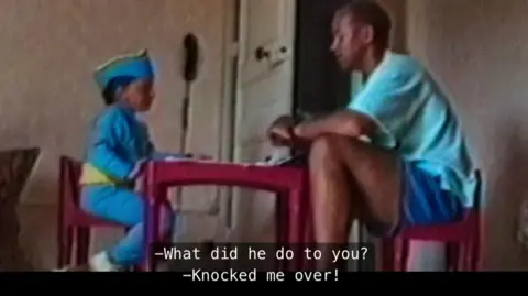 The Murder of Rachel Nickell, Netflix A small child and an adult sitting at a tiny red table indoors, with subtitles on screen reading, “What did he do to you? – Knocked me over!”. Both dad and son wear blue.