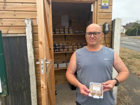 Julian Manser stands outside the Capel Cake Shed with shelves of packaged cakes visible behind him.
