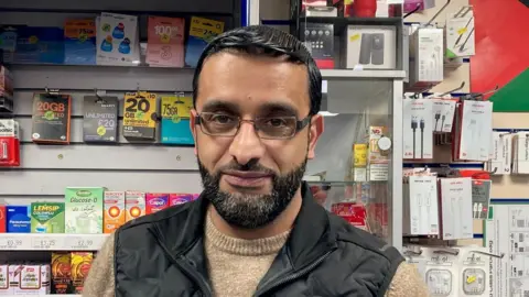 A man wearing glasses and a black gilet and light brown jumper with shop shelves in the background 