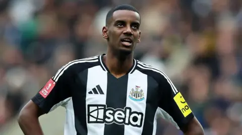 Alexander Isak stands with his hands on his hips in a football stadium looking towards the sky. He is wearing a black and white Newcastle shirt.