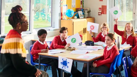 Generic image of a classroom, with primary aged pupils seated at a table holding up pieces of paper with ticks and crosses on them, while a teacher is on the left