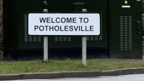 Paula Cornford-Cousins A road sign which has been defaced and reads: "Welcome to Potholesville". There is a green cabinet behind the sign.