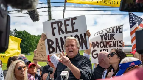 Getty Images Steve Bannon stands in front of a microphone speaking to a crowd, while someone holds a "free Bannon" sign behind his head. He has grey hair and is earing a black shirt.