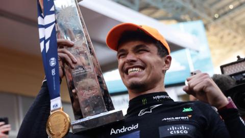 Lando Norris of McLaren celebrates with the team after the Formula 1 Miami Grand Prix at Miami International Autodrome in Miami, United States on May 5, 2024. Lando is a 24-year-old white man with short brown hair. He grins, holding his trophy and gold medal. He wears his black racing gear covered in sponsors' logos and an orange cap.