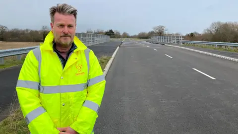 Councillor Chris Watts is wearing a hi-vis, long sleeve jacket. He is stood on the side of the empty road, which has pavements on either side of the carriageway.