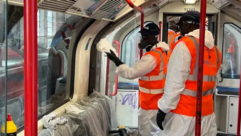 Two men dressed in white protective overalls and hi viz vests clean the graffiti inside a train with buffers