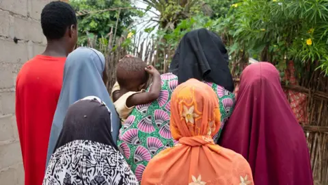 BBC Fatima, dengan jubah bermotif hijau dan merah muda, menggendong anaknya yang berusia dua tahun dengan kaus kuning. Dia dikelilingi oleh lima anaknya yang lain - empat gadis mengenakan kepala berwarna berbeda dan seorang remaja putra dengan kaus merah. Mereka semua memiliki punggung ke kamera sehingga Anda tidak dapat melihat wajah mereka.