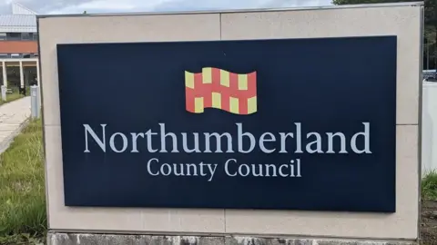 A large navy blue Northumberland County Council sign which has white lettering. Above the words is a Northumberland flag which has yellow and red squares. There is grass behind the sign and a brick building.