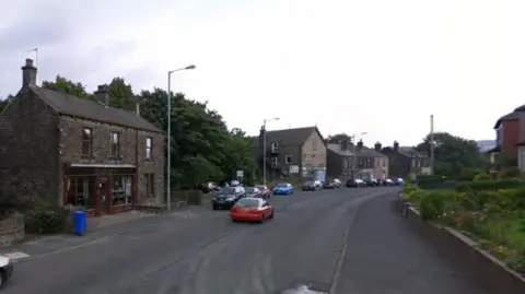A street view image of a residential area with aged stone buildings hugging the road on the left. Several cars are parked along the curb, with a red car travelling on the left side of the road.