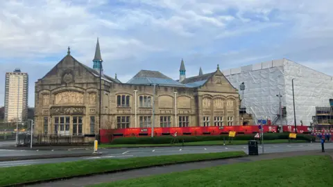 A two-storey stone historical building seen from across a roadway, with parts of the building cordoned off with fencing, while yellow signs warn vehicles approaching the site.