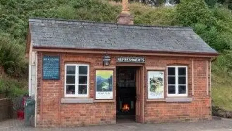 Jason Hood/SVR A close-up of a small brick rail station, which has signs and posters on it and a grass verge behind it. 