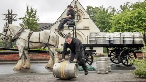 PA Media A shire horse on the left of the picture with a horse drawn cart with seven silver barrels on top of the cart. A man is seen sitting on top of the cart wearing a hat and a second man is in front of the horse and cart rolling a barrel. They are pictured in front of a white walled church with trees and bushes to the right of the picture and a wall to the left