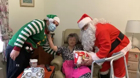 Irene Chantry sits in an armchair and pose with care home staff. One person, to her left, is dressed in a green elf's costume and green Christmas hat. Another person, to her right, is dressed in a Father Christmas costume.