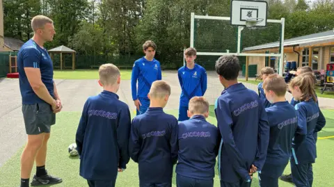 A group of eight children line up in football kits opposite two older teenage boys while an adult male teacher looks at them.