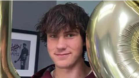 A close-up of a man with brown hair smiling at the camera. He is holding gold balloons either side and there is a small framed photo in the background. 