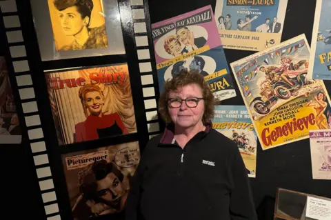 A woman standing in front of a display of 1950s film posters and magazine covers, all showing the actress Kay Kendall. They include posters for the film Genevieve and magazines called True Story and Picturegoer. The woman has curly brown hair and is wearing glasses and a black fleece.